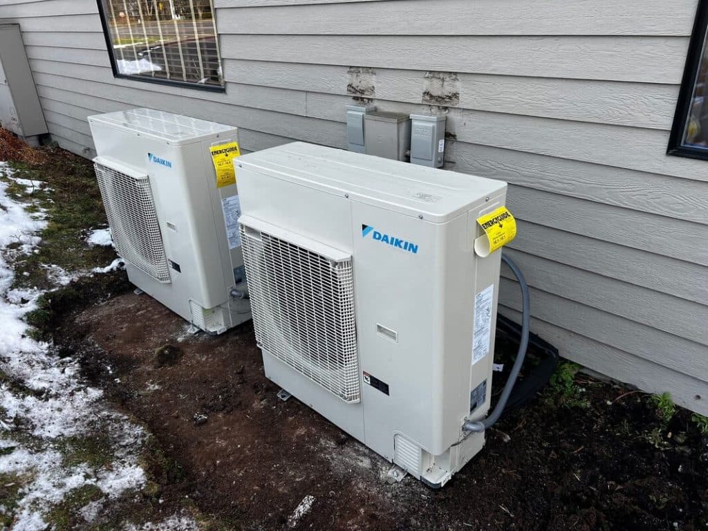 A white Daikin heat pump condenser newly installed on a gravel pad beside grey lap siding, lines run neatly to the wall.
