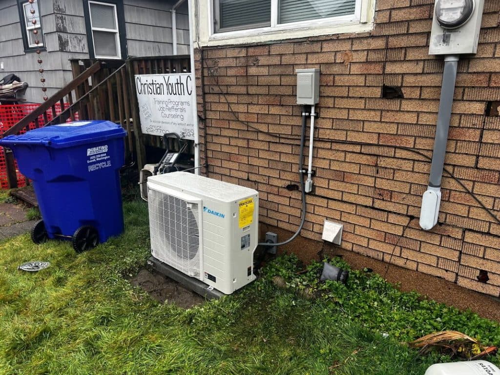 An outdoor condenser unit on its pad against a PNW home, the surrounding ground still wet from rain.